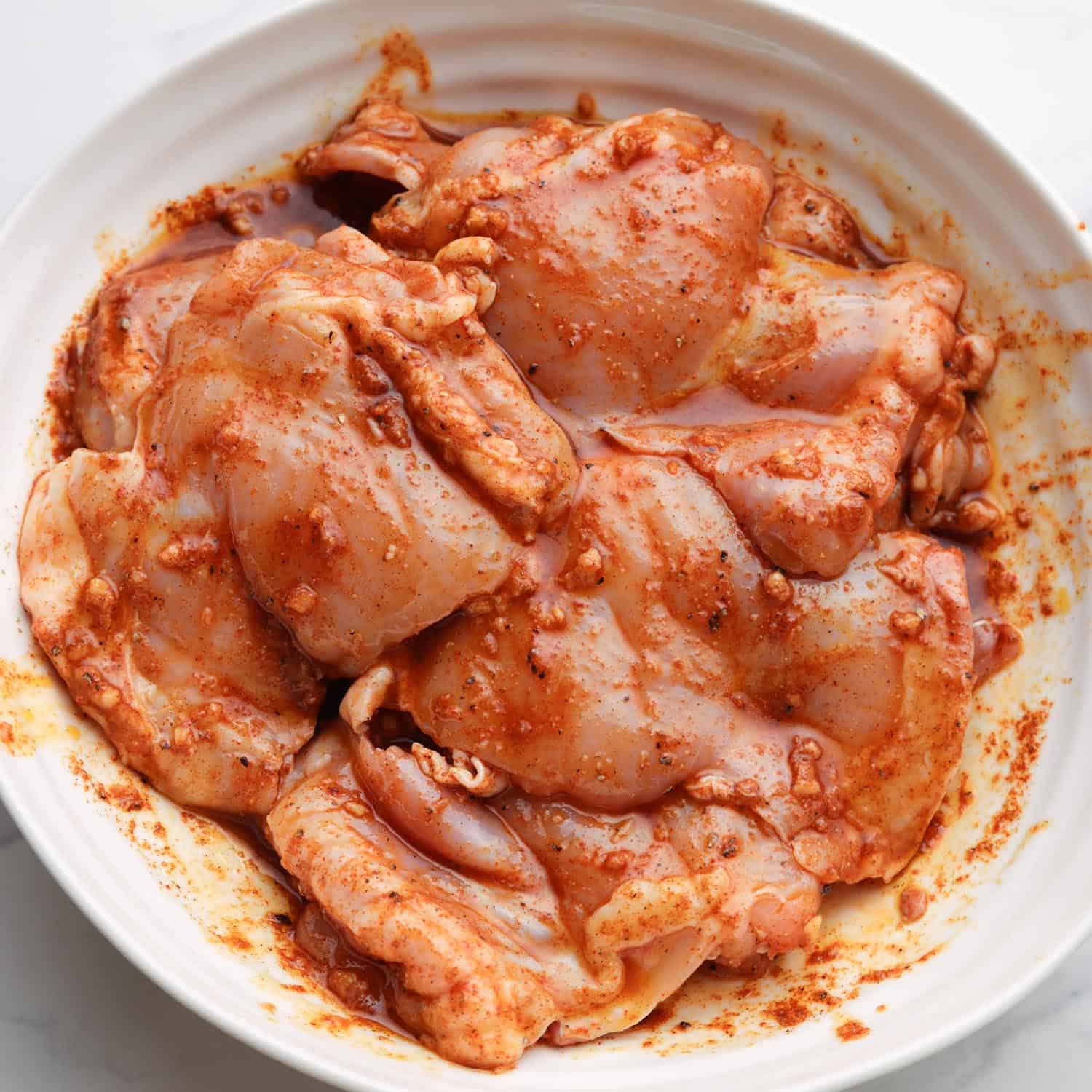 chicken thighs marinating in a red garlic and spice blend in a white bowl. 