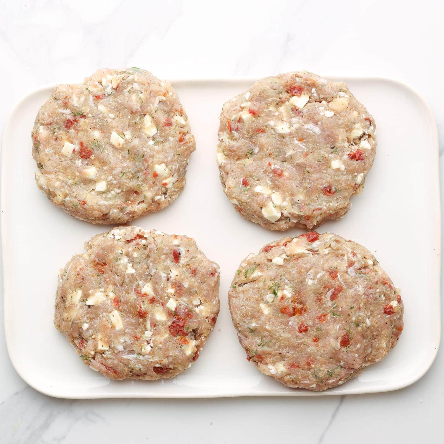 greek chicken burgers with feta cheese and sundried tomatoes on a white rectangular platter.