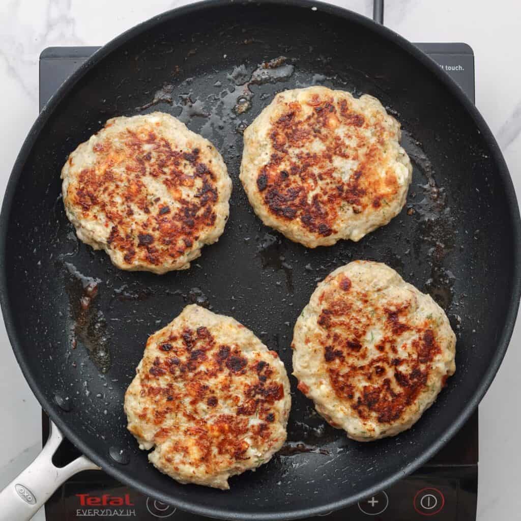 four greek chicken burgers cooking in a nonstick skillet.