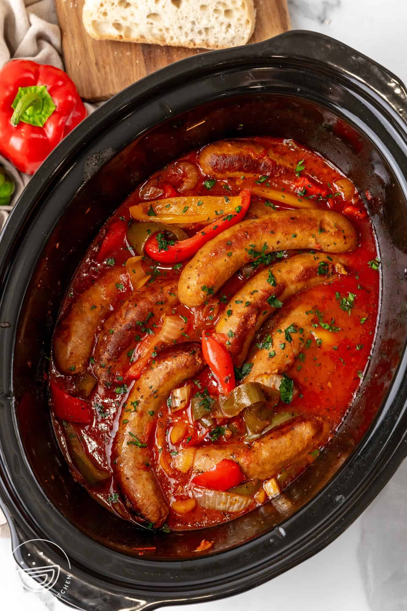 overhead view of a black oval crockpot filled with cooked italian sausages and peppers in a tomato sauce.