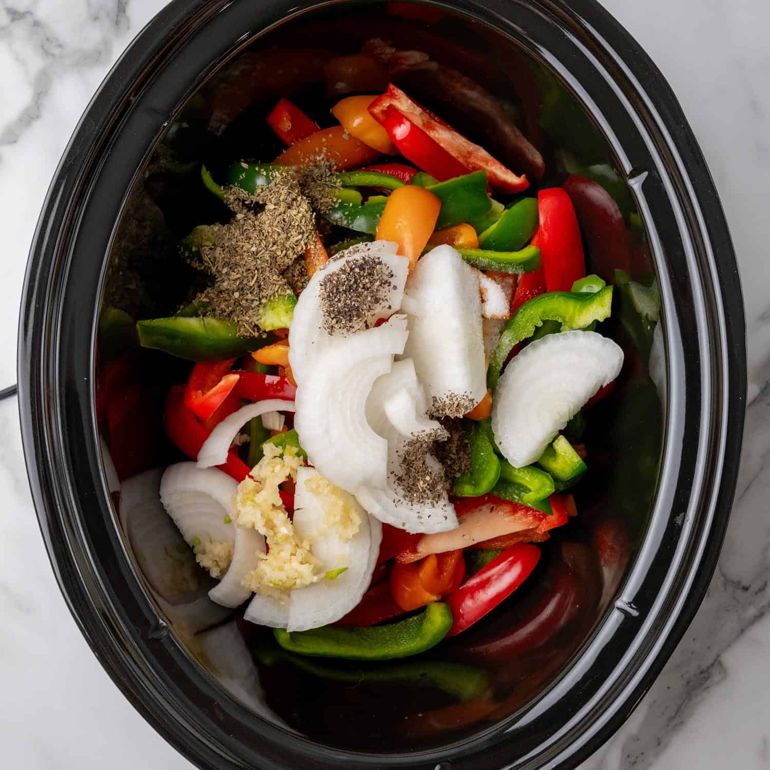 sliced bell peppers and onions in a black crock pot, sprinkled with seasonings.