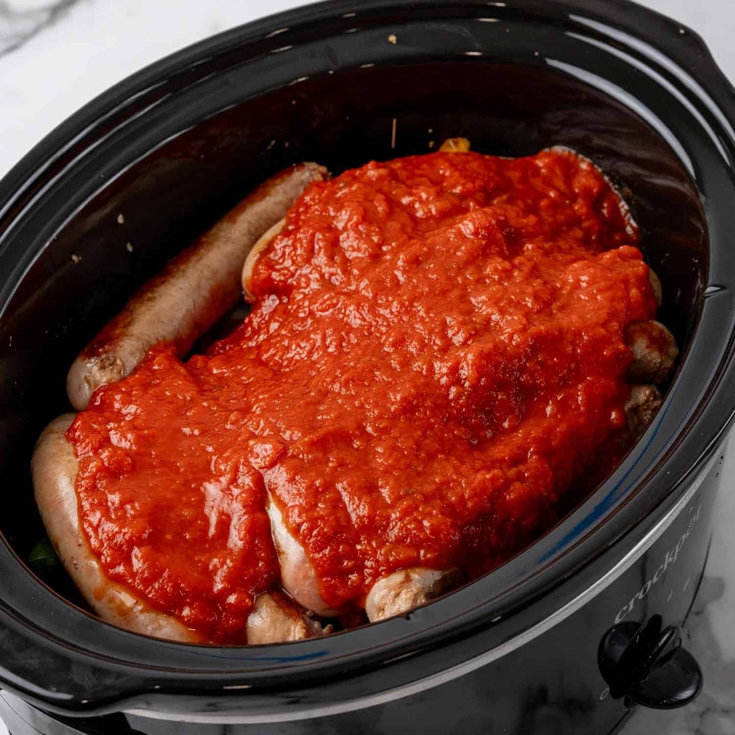 marinara sauce poured over sausage links in a black oval crock pot.