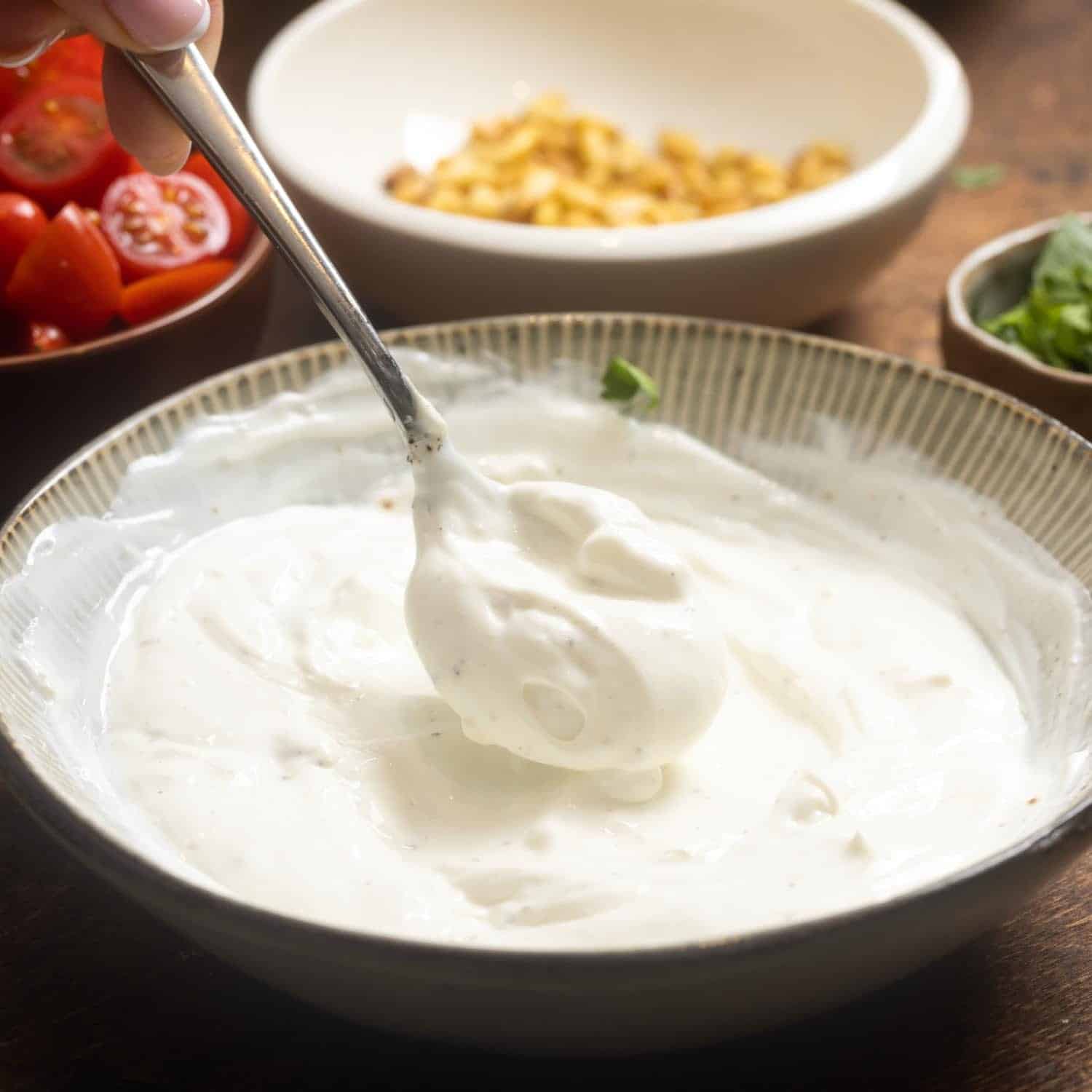 Garlicy yogurt for turkish pasta in a ceramic bowl. In the background is a bowl of chopped tomatoes and a bowl of toasted pine nuts.