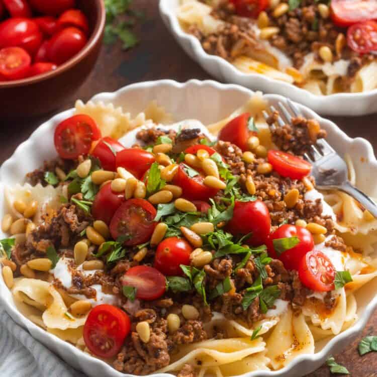 Two bowls of Turkish pasta topped with yogurt, ground beef, tomatoes, pinenuts, and herbs.