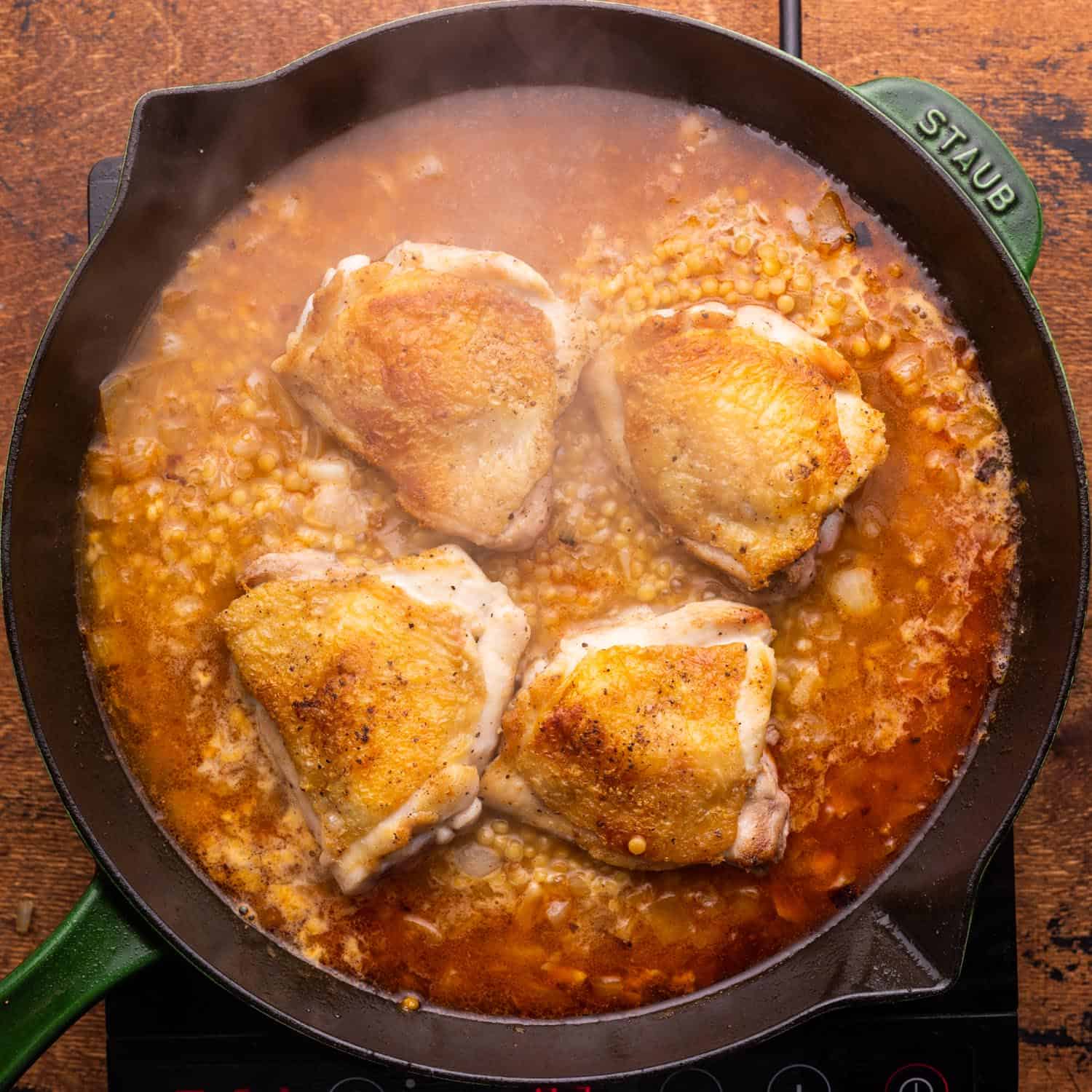 seared chicken thighs on top of couscous cooking in broth, inside of a cast iron skillet.