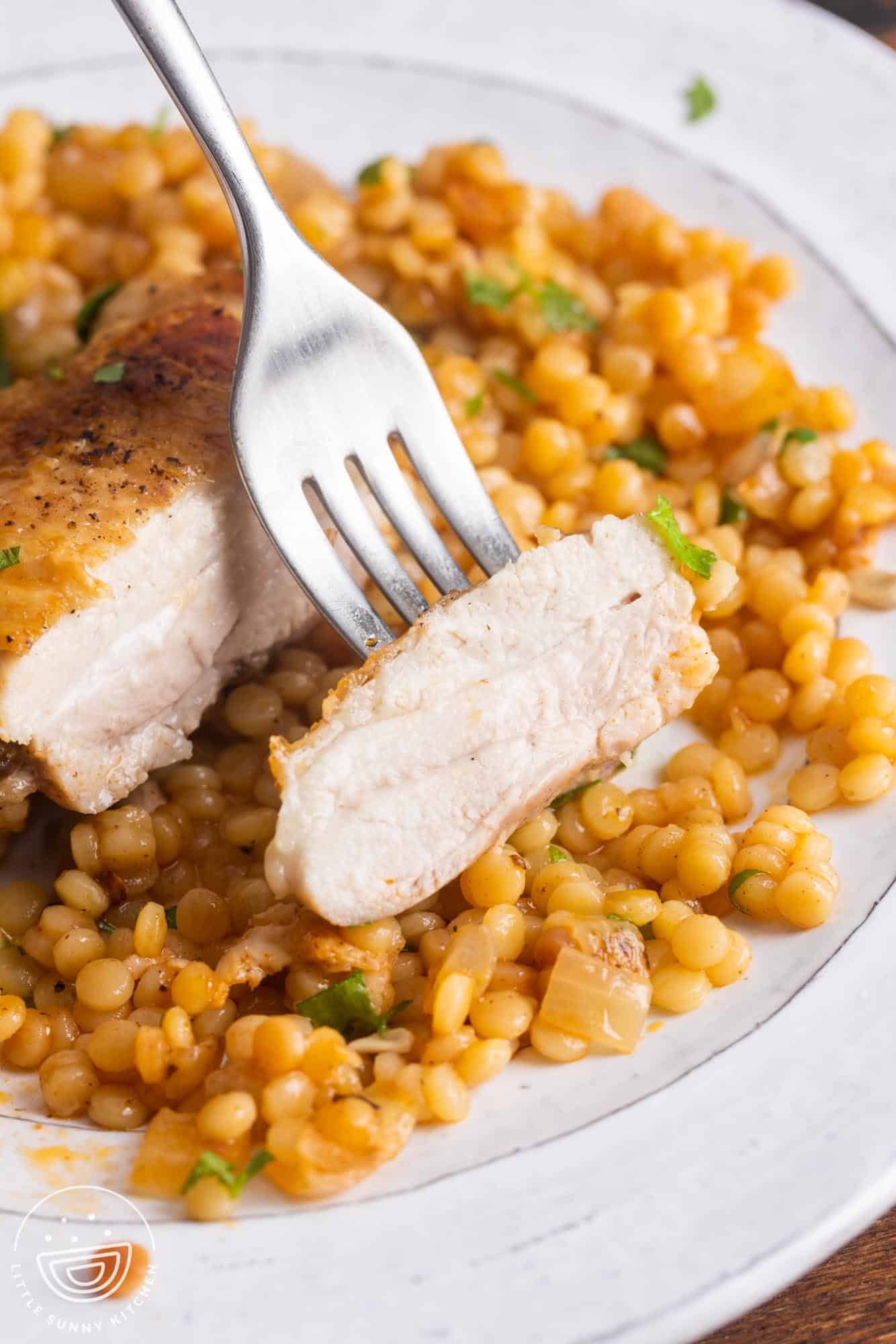 chicken and couscous on a white plate. A fork is picking up a bite of chicken to show the juicy meat.