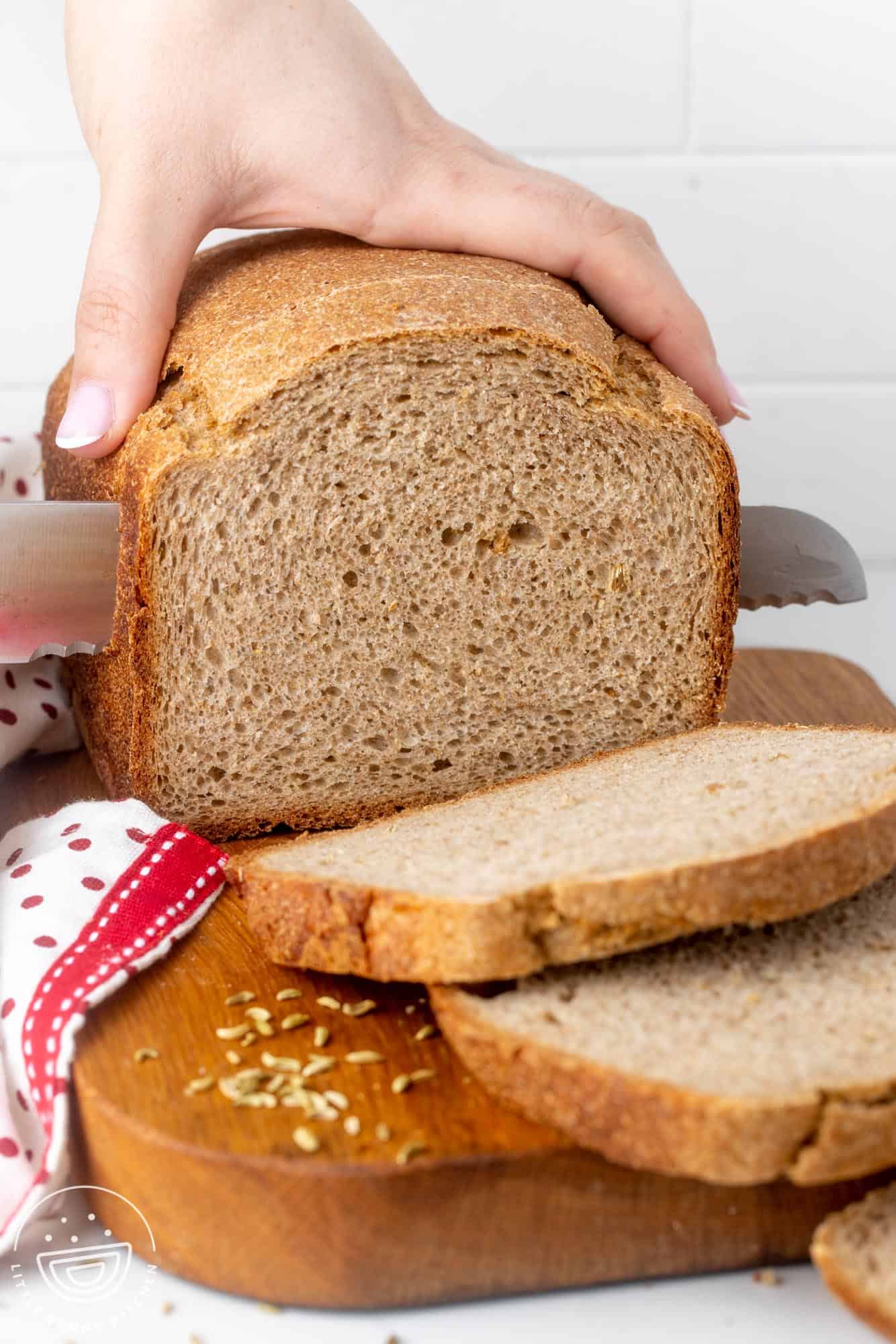 a loaf of rye bread on a wooden board. A hand is holding the loaf and cutting it into slices with a large bread knife.