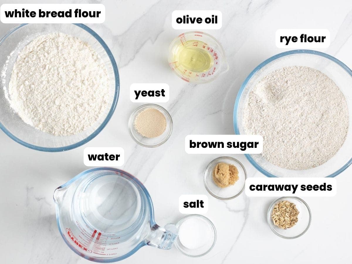 Ingredients for making rye bread in a bread machine, including brown sugar and caraway seeds, all in separate glass bowls, arranged on a marble surface.