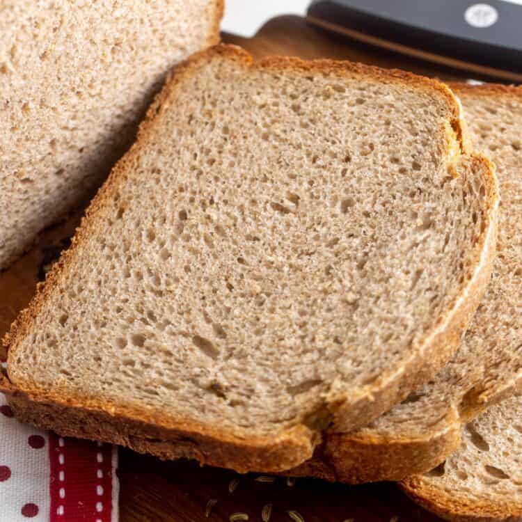 slices of bread machine rye bread on a wooden cutting board. Caraway seeds are sprinkled on the board as well.