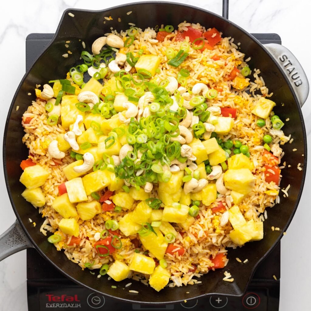 Pineapple, cashews, peas, and green onion added to fried rice in a large skillet.