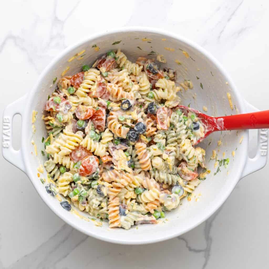 bacon ranch pasta salad stirred in a large white mixing bowl with a red spoon.