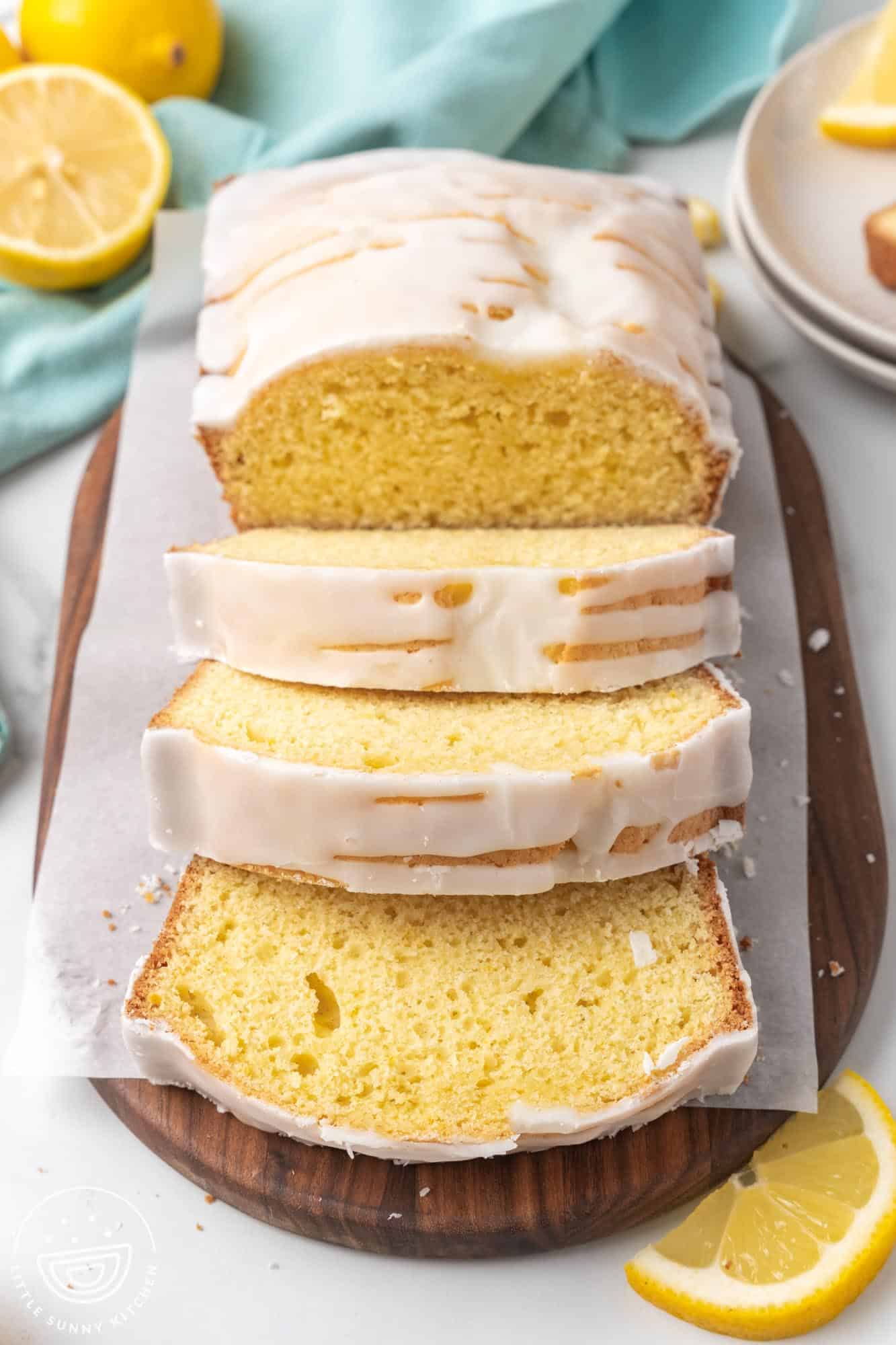 A sliced lemon loaf cake with a white glaze, displayed on a wooden cutting board. The cake has a soft, yellow crumb and is surrounded by fresh lemon wedges and a light teal cloth in the background.