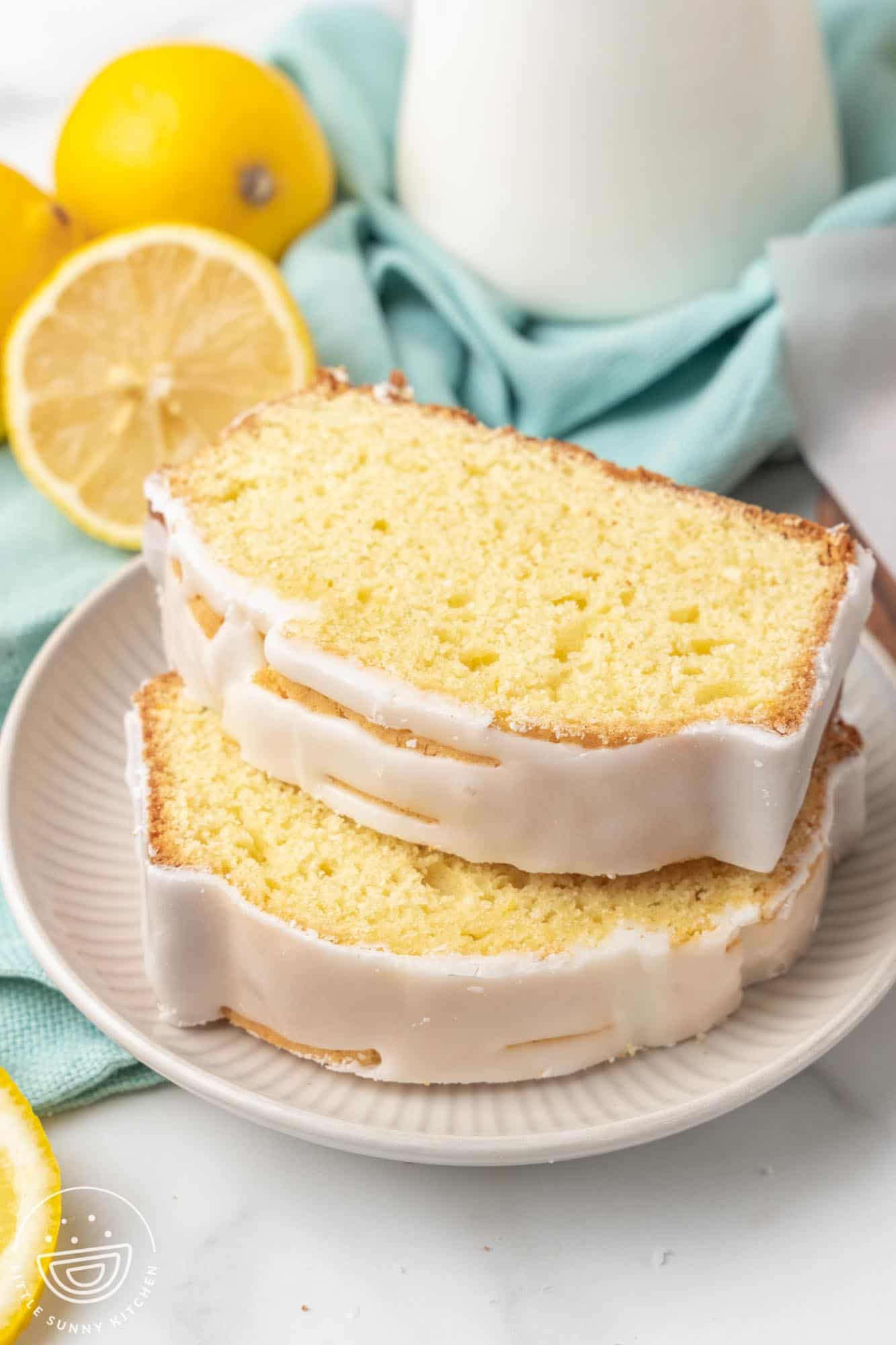 Two slices of lemon loaf cake with a white glaze stacked on a plate. The background features whole lemons, a teal cloth, and a milk jug, adding a fresh and cozy atmosphere.