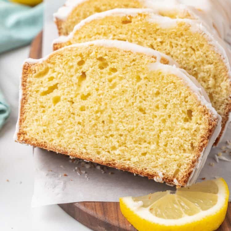 Close-up of three slices of lemon loaf cake with white glaze arranged on a wooden cutting board. A lemon wedge sits next to the cake, and crumbs are scattered on the parchment paper beneath it.