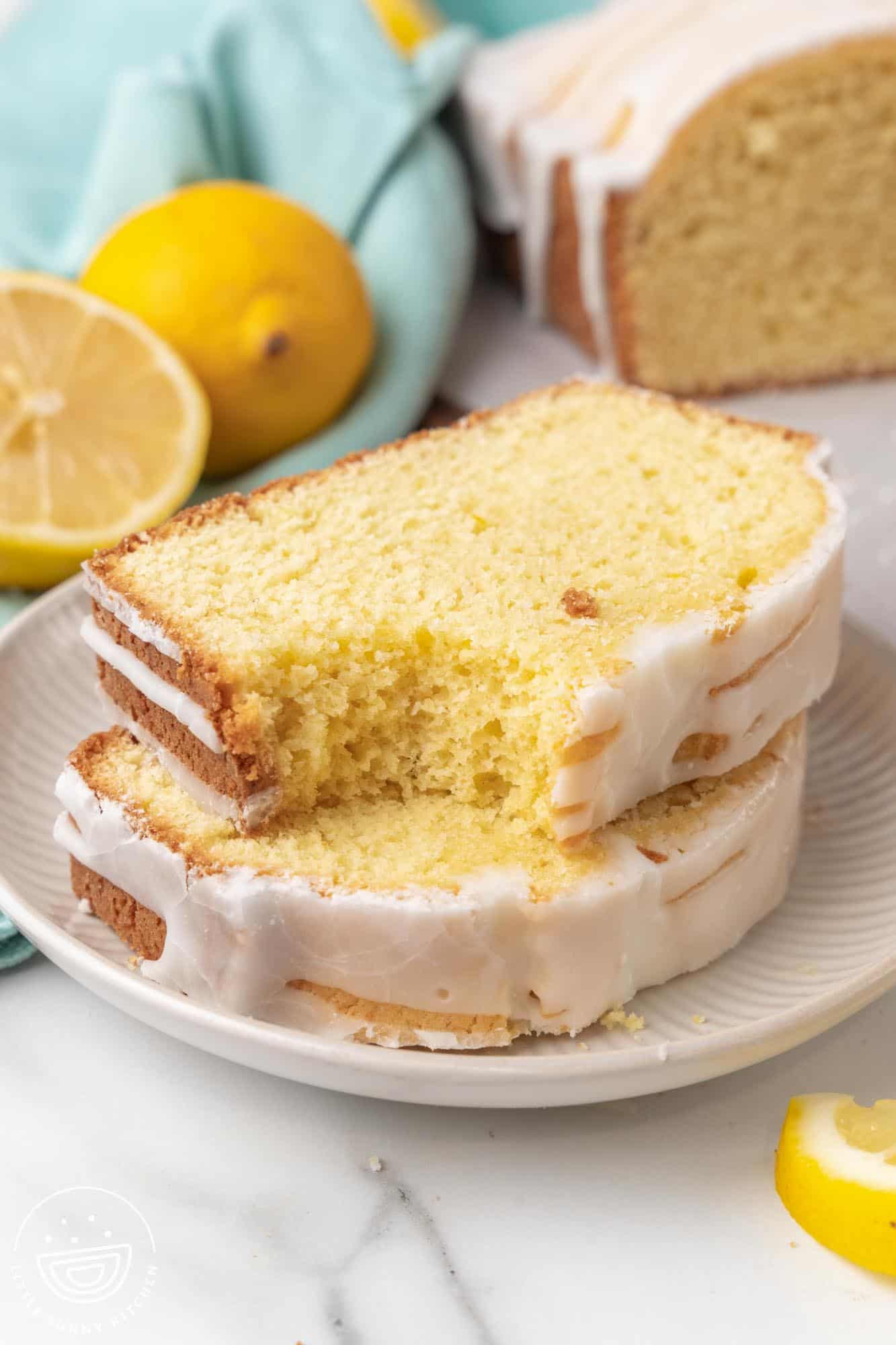 Two slices of lemon loaf cake stacked on a white plate, with a bite taken out of the top slice. The plate is set on a marble surface with whole and halved lemons in the background, along with a light teal cloth.