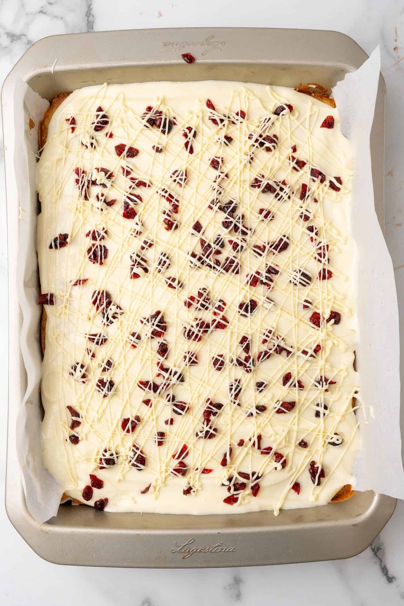 cranberry bliss bars in the pan, covered with frosting and dried cranberries, drizzled with lines of white chocolate.