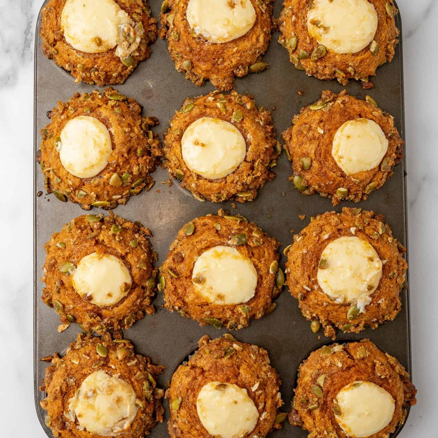 pumpkin cream cheese muffins baked in a muffin tin. The muffins are filled with cream cheese and topped with pepitas.